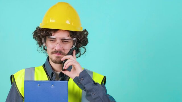 Handsome young construction worker with safety glasses and helmet that is thinking while holding clipboard and talking on mobile phone against turquoise background.