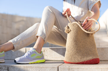 Unrecognizable young woman in light clothes sitting on concrete stairs with handmade knitted bag. Eco-friendly shopping. Zero waste trend. DIY jute bag