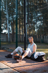 Fototapeta na wymiar Basketball player cool young guy having break after training sitting with phone on court outdoors in summer