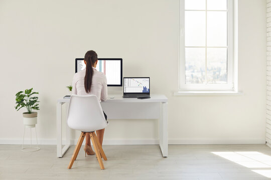 Back view of a woman working in the interior of a clean spacious office. Young female worker working with spreadsheets sitting at a desk with a modern desktop computer and a laptop
