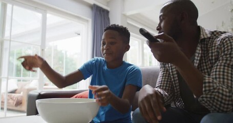 Happy african american father and excited son watching tv and eating popcorn in living room - Powered by Adobe