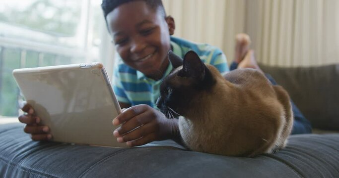 Happy african american boy lying on couch using tablet,stroking siamese pet cat and smiling