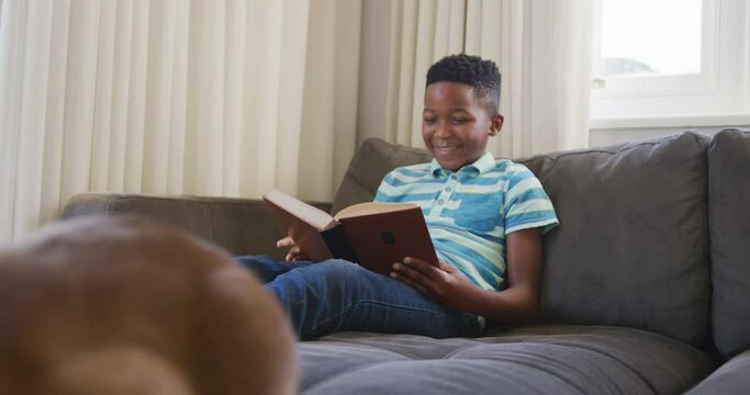 Happy african american boy sitting on couch reading book and smiling, pet cat sitting in foreground