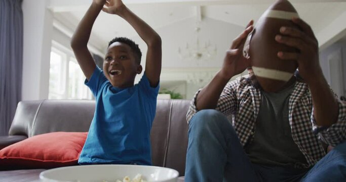 Excited african american father and son watching football on tv, cheering and high fiving