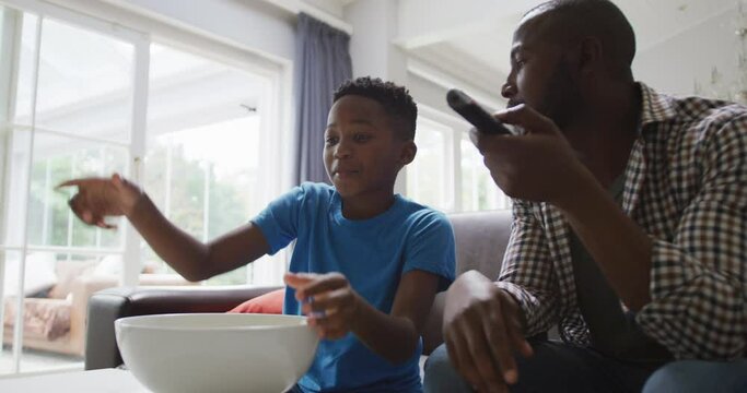 Happy african american father and excited son watching tv and eating popcorn in living room