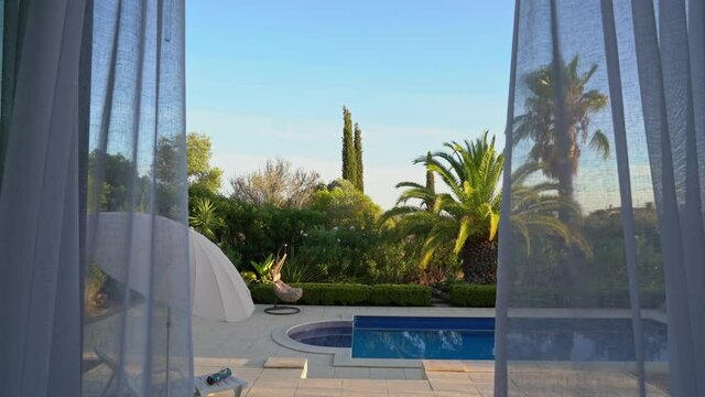View from modern living room with curtains blowing in the wind revealing a balcony and an outside pool. And tropical garden.