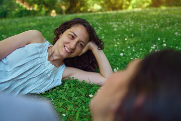 Young woman looks and smiles at her out of focus partner. She is happy spending time with her girlfriend, lying on the grass.