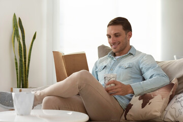 Young man sitting on a sofa reading a book while having coffee at home, lifestyle, studying