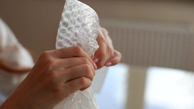 Close-up hands of unrecognizable young woman playing pops bubble wrap to calm herself sitting at table. Closeup view of female popping plastic bubblewrap at home office. Shooting in slow motion.