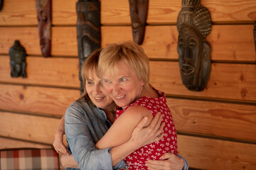 Two women 55 years old girlfriends hugging against the background of wooden wall, posing for a photographer