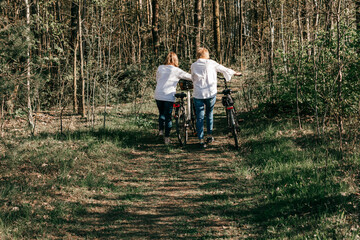 Two female mature friends are walking along the road with bicycles in forest. Back view