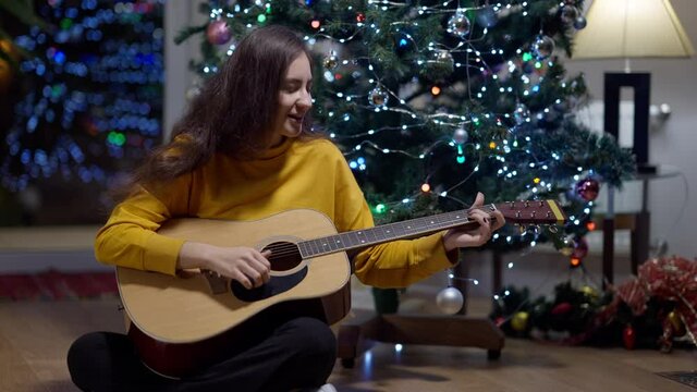 Wide shot of confident cheerful Middle Eastern young woman playing guitar singing sitting at decorated Christmas tree on New Year's eve. Portrait of talented female musician celebrating holiday at