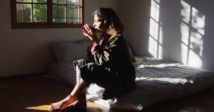 Relaxed mixed race woman wearing headphones sitting on bed drinking tea in sunny cottage bedroom