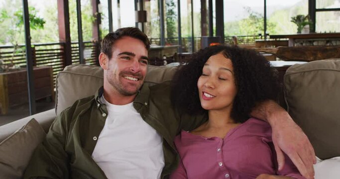 Happy diverse couple sitting on couch in living room, watching tv, embracing and talking