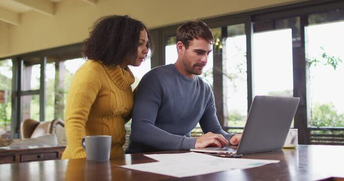 Diverse couple talking in living room, using laptop, looking at paperwork and paying bills