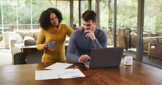 Happy diverse couple talking in living room, using laptop, looking at paperwork and paying bills