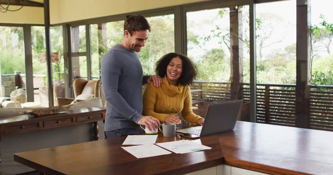 Diverse Couple Talking In Living Room, Using Laptop, Looking At Paperwork And Paying Bills