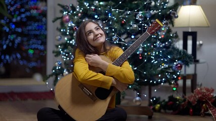 Young talented Middle Eastern woman dreaming sitting with guitar at home on Christmas eve. Portrait of confident happy beautiful musician enjoying New Year holiday indoors in winter