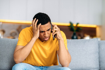 Confused sad latino guy sitting on a sofa in the living room, dressed in casual yellow t-shirt, bewildered, talking on the phone, angry, annoyed, have unpleasant telephone conversation