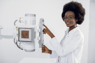 Beautiful African woman in white medical coat, standing near the X-Ray machine in modern hospital...