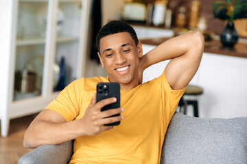 Satisfied joyful hispanic guy dressed in a yellow t-shirt, sitting in living room on a sofa,...
