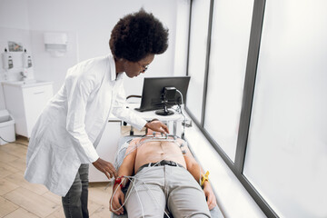 ECG test of young man at modern clinic. Young African lady doctor cardiologist putting special...