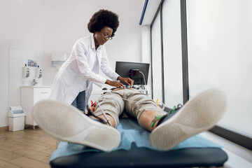Heart electrocardiogram for young man patient. Young African American doctor attaches vacuum sensors on male patient for recording his electrocardiogram in modern light clinic