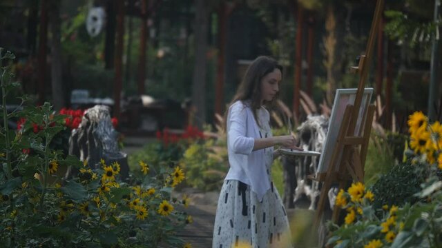 Girl artist in a white dress at work in the flower garden. Doing oil painting on canvas. With loose hair, concentrated, focused, hard-working and professional.