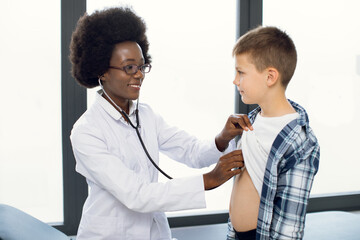 Cute likable Caucasian teen boy and joyful African woman doctor, during doctors checkup. Pediatrist examinates young patient's heartbeat and lungs with stethoscope
