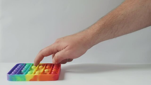 Male hand playing with a rainbow Pop It Toy on a white background. Raises his thumb up after the game. Fashionable colorful anti-stress fidget bubble popping toy. Copy space, close up