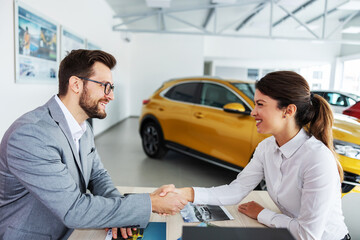 Smiling, friendly car seller sitting at the table with a customer and shaking hands. Woman just...
