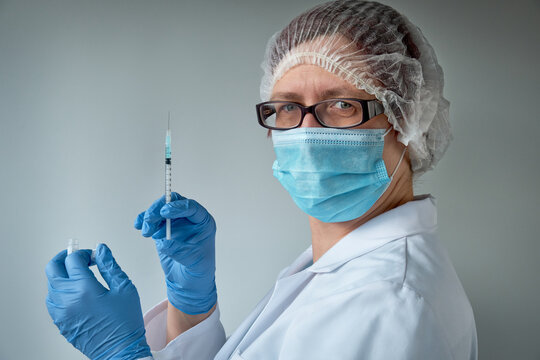 Medic, doctor or nurse with covid-19 vaccine vial and syringe. Mature Caucasian, European female practitioner in face mask, glasses and disposable hat, blue nitril gloves and protective white gown.