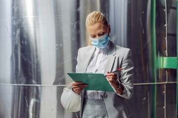 Dedicated successful blond female supervisor in formal wear with protective face mask filling up documentation while standing in power plant during corona.