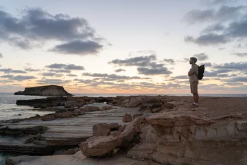 Gordijnen Sea sunset view. Man with backpack on rocks with beautiful view of Yeronisos Island near coast of Agios Georgios Pegeias. Guy enjoying ocean horizon, panoramic sunset in cyprus on mediterranean sea © Elizaveta