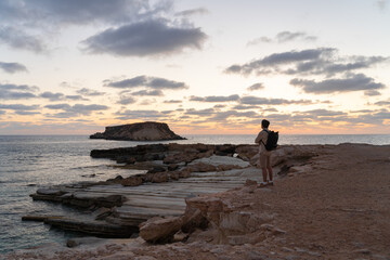 Sea sunset view. Man with backpack on rocks with beautiful view of Yeronisos Island near coast of Agios Georgios Pegeias. Guy enjoying ocean horizon, panoramic sunset in cyprus on mediterranean sea