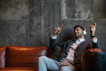 A businessman sitting on the couch in front of a gray wall with headphones on and enjoying the...