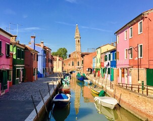 Fototapeta na wymiar A beautiful view looking down a canal in Burano, Italy with the leaning tower in the background. This popular island is full of houses of all different colours