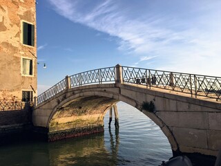 An old small historic stone bridge with iron railings crossing a canal with the open ocean in the background in Northern Venice on a beautiful summer morning.