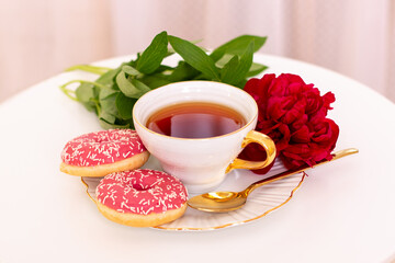 Cup of tea, sweet strawberry donuts on golden plate with peony flower