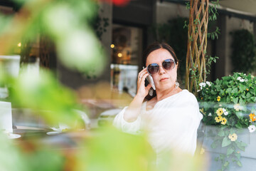 Beautiful brunette middle aged woman in sunglasses in white clothes talking on mobile phone in hand...