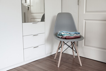 Stack of folded clean clothes on modern chair beside wardrobe in the bedroom.