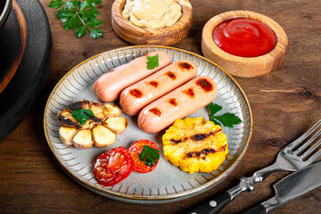  Grilled sausages with vegetables and sauces. Summer BBQ with sausages, corn and tomatoes on the wooden table close up