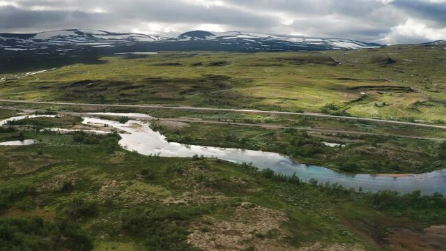 A glimpse on the tranquil nature of the Arctic circle: a shallow river is calmly running near a railway,  an asphalt road is neighboring with green hillside, snowy peaks are touching cloudy sky.