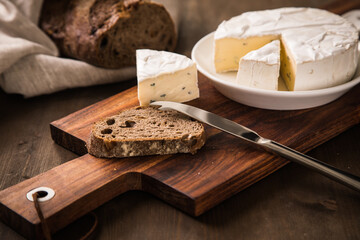Loaf of soft blue cheese from cow milk on porcelain plate with walnut bread, knife, linen towel and dark brown wooden board as snack or dinner