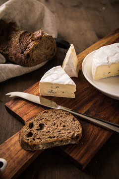 Loaf of soft blue cheese from cow milk on porcelain plate with walnut bread, knife, linen towel and dark brown wooden board as snack or dinner