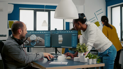 Multiethnic team discussing about industrial project using dual monitors setup to desing 3D metalic pieces in CAD software. Industrial engineer and african manager working together in creative office