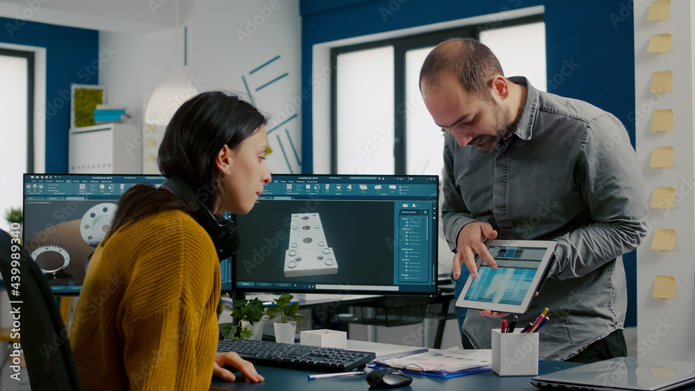 Wall mural Industrial designer discussing with colleague holding tablet with in CAD program, designing 3D prototype of building. Woman engineer using pc with two monitors sharing ideas for construction mechanism