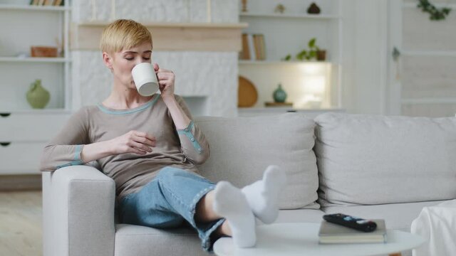 Dreamy carefree happy woman sitting on sofa drinking morning coffee or warm tea at home, smiling attractive lady holding cup relaxing feels stress-free enjoying cozy lifestyle on pleasant weekend