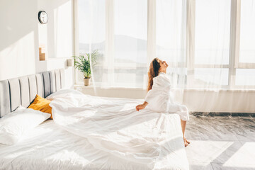 Tranquil young woman in white bathrobe sitting on large comfortable bed near panoramic window in hotel room at sunrise.