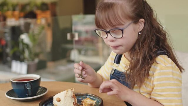 Over-the-shoulder waist-up shot of pretty 11-year-old girl with down syndrome in eyeglasses eating cake in cafe looking at caring mother removing crumbs from daughter face